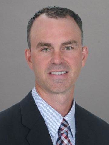Image of Karl Kurt dressed in a black suit, blue checkered shirt and multi-colored tie.